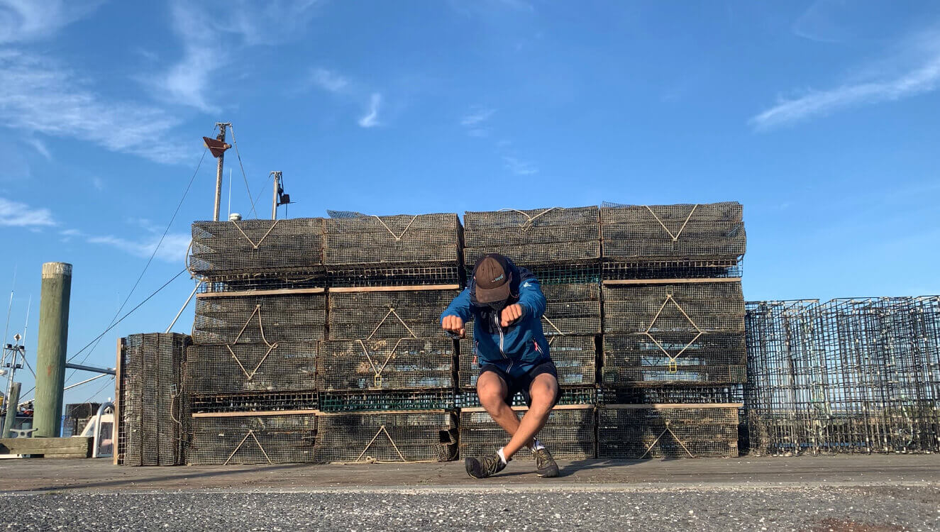 L'interprète en danse Guillaume Loslier-Pinard, est devant une pile de caisses de homards vides, dans une posture dynamique, poings vers l'avant, jambes fléchies et croisées