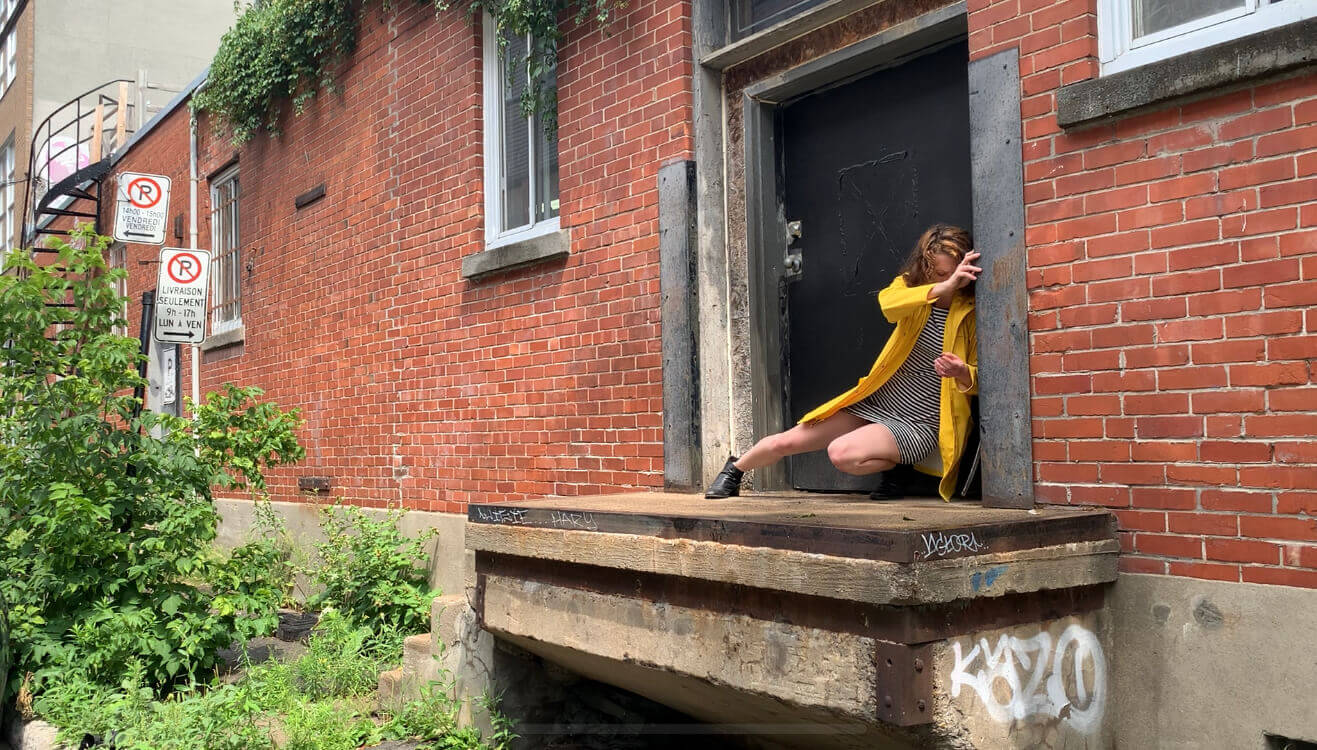 L'interprète en danse Louise Gamain, dans un imperméable jaune, s'appuie contre un cadre de porte, à l'extérieur.